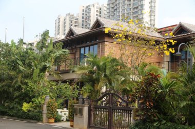 Çit kapısının önündeki yemyeşil ve tropikal bitkilerle çevrili iki katlı bir villa, yüksek binalar ve modern mimarinin yer aldığı kentsel bir zemin üzerine kurulmuş.