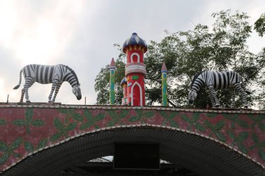 Hint temalı park girişi, kapının üstünde zebra heykelleri, Hint boyalı bir duvar, ve önünde kemerler. Renk paleti kırmızı, beyaz ve yeşildir. Merkezde renkli bir kule duruyor..