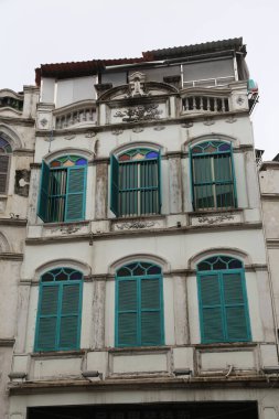 Mavi panjurlu ve beyaz çatılı yüksek bir bina. Binanın kemerli pencereleri ve balkonu olan eşsiz bir tasarımı var.