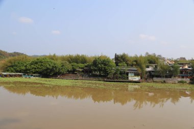 Diğer tarafında bir sürü ağaç ve ev olan bir nehir. Su kahverengidir. Suda tekneler var.