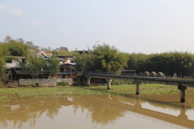 Güney Çin 'in kırsal kesiminde, yanında ağaçlar ve binalar olan bir nehir kıyısında küçük bir köprü. Su kahverengimsi yeşil, yukarıdan yansıtan ışık.