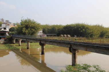 Kırsal kesimlerdeki nehir üzerindeki bir köprü, eski bir trenin geçtiği beton ve demir korkuluklardan oluşur. Yolun her iki tarafında yeşil bitki örtüsü, bambu ormanları, çiftlik evleri ve diğer binalar var.
