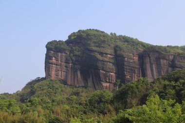 Tepesi yeşil bitki örtüsü ve ağaçlarla kaplı, güney Çin 'deki diğer dağ zirveleriyle çevrili dev bir kaya duvarı..