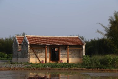 Güney Çin 'in kırsal kesimlerinde yerel malzemelerden yapılmış ve çatısında kırmızı fayanslar bulunan küçük bir ev. Bina, çevresinde sulak bitki örtüsü bulunan eski bir nehrin yanında yer alıyor..