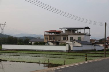 Uzakta, Çin 'in kırsal bir bölgesinde beyaz duvarları ve kırmızı kiremitleri olan iki katlı bir bina var. Evin pencerelerinin üzerinde küçük bir çatısı var..