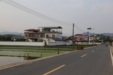 Kameranın önünde, her iki tarafında evler ve pirinç tarlaları olan bir yol. Bir evin yanında güneş panelleri olan bir alan var..