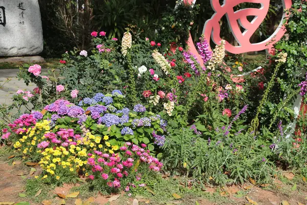 Arka planda pembe işareti olan bir bahçe. Tabelada bir üçgen var. Bahçede bir sürü çiçek ve bitki var.