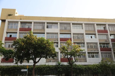 İki katlı öğrenci yurdunun köşesi, kırmızı ve beyaz fayanslı düz bir cephe duvarı, tüm katlarda dikdörtgen pencereler, alt katta beton bir bina, önünde büyük ağaçlar.