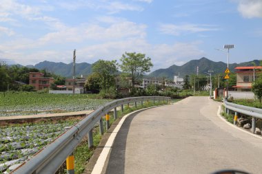 Asfalt yol köye çıkıyor. Uzakta yeşil dağlar ve bir tarafında elektrik depolama sistemi var. Yakındaki sebze tarlalarında nilüfer çiçekleri açıyor..