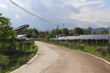 Asfalt bir yolun kenarında yeşil bitki örtüsü ve kırsal alanlarda alçak beton duvarlarla çevrili bir güneş enerjisi santrali bulunmaktadır. Yol yüzeyi üzerine monte edilmiş güneş panellerinden gelen güneş ışığını yansıtır..