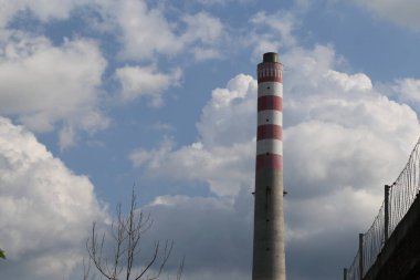 Endüstriyel bir binanın sağ tarafında bulutlu bir gökyüzüne karşı büyük beyaz ve kırmızı çizgili bir baca. Arkasından çıkan duman, siluet ya da figürlere benzeyen bulutlar oluşturur.