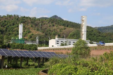 Yeşil dağların ve mavi gökyüzünün arka planında beyaz duvarları ve gri kiremitleri olan bir endüstriyel fabrika binası var. Önünde iki büyük güneş paneli var, her iki tarafında.