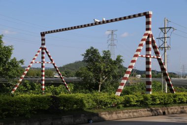 İki kırmızı ve beyaz çizgili A biçimli çelik çerçeve heykel, yeşil ağaçlar ve mavi gökyüzü ile endüstriyel bir alanın arka planına kurulmuş. Heykeller, yüksek hızlı rayların bir tarafında birbirlerine dik açıda duruyorlar.