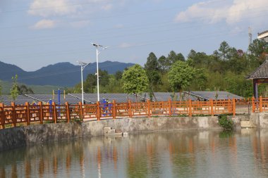 Parkta, üzerinde güneş panelleri olan turuncu ahşap bir korkuluk köprüsü ve arkasında yeşil ağaçlar var. Kırsal bölgelerde gölün önünde çeşitli çiçeklerle süslenmiş küçük bir meydan vardı..