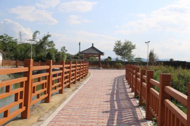 Kırmızı tuğlalı bir yol balık havuzuna gidiyor, her iki tarafında da tahta korkuluklar var. Çin mimarisi tarzında antik bir pavyon yakınlarda duruyor. Arkaplanda yeşil ağaçlar ve mavi bir gökyüzü var..