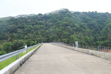 Asfalt yol yeşil dağlar ve ormanlarla çevrili ve sağ tarafında demir korkuluklar var. Bir uçtaki beton baraj, yemyeşil ağaçların ve gökyüzünün arka planında beyaz ışıkta parlıyor..