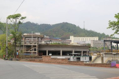 Öndeki caddenin altındaki bir yeraltı otoparkının büyük bir inşaat alanı, yanında iki katlı ve birkaç yüksek konteynırlı bir bina bulunuyor. Arka planda ormanla kaplı tepeleri görebilirsiniz..