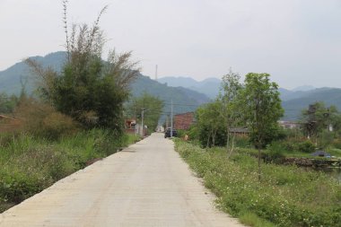 Köye giden uzun beton bir yol var, her iki tarafında yeşil bitki örtüsü ve arkasında dağlar var. Caddenin bir ucunda küçük ağaçlarla çevrili eski bir kırmızı tuğla ev var.