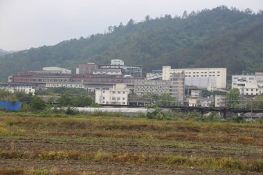 Uzakta, önünde bir sanayi parkı ve her iki tarafında beyaz binalar var. Yakın mesafede, içinden yeşil bitkiler çıkan kahverengi toprak görüyoruz..