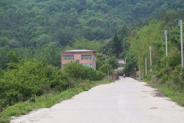 Uzakta, Çin kırsalında iki toprak yolun bir tarafında eski bir bina var, bu yolların her iki tarafında yeşil ağaçlar ve çalılar büyüyor..