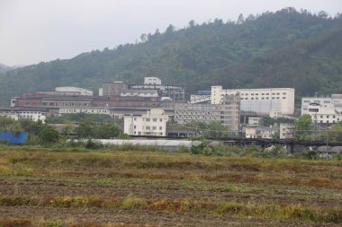 Uzakta, önünde bir sanayi parkı ve her iki tarafında beyaz binalar var. Yakın mesafede, içinden yeşil bitkiler çıkan kahverengi toprak görüyoruz..