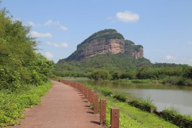 Danxia Dağı 'nın eteklerine giden kırmızı tuğla yol yemyeşil bitki örtüsüyle kaplı ve önünde gökyüzüne doğru yükselen yüksek bir dağ zirvesi var. Nehir sessizce akar.
