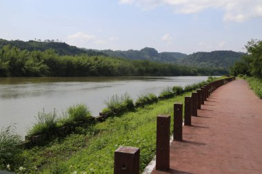 Kırmızı tuğla yol, yeşil bitki ve ağaçların her iki tarafındaki nehir boyunca uzanan ahşap bariyerlerle kaplı. Önünde, mavi gökyüzünün altında sonsuz bir su yüzeyi görebilirsiniz..