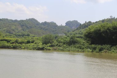 Arka planda orman olan bir su kütlesi. Su kahverengi ve bulanık. Uzakta bir ev var.