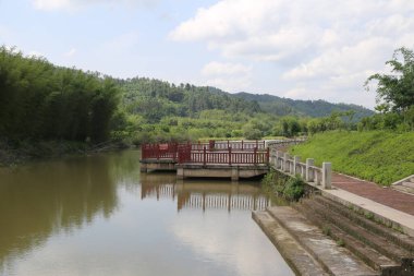 Çin mimarisi tarzında terk edilmiş bir köprüye sahip nehir kıyısının geniş bir manzarası, her iki tarafında yemyeşil bitki örtüsü ve tepelerle çevrili. Su gökyüzünü yansıtır, uzak bulutların yansımalarını yaratır..