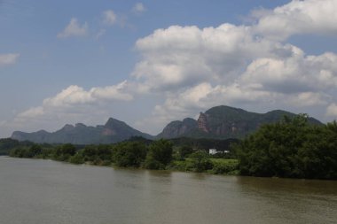 Nehir, her iki tarafında dalgalanan dağlar ve kırmızı uçurumlar bulunan yeşil bitki örtüsüyle çevrilidir. Gökyüzünde süzülen beyaz bulutlar var..