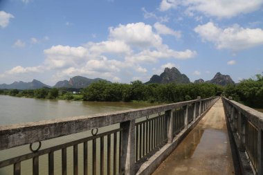 Nehir boyunca uzanan taş köprü yeşil ağaçlarla kaplı ve önünde bulutlu mavi bir gökyüzünün altında sonsuz bir su yüzeyi uzanıyor. Çin 'de nehrin iki yakasında da dağlar vardı..