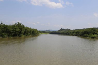 Nehir, her iki tarafında yeşil bitki örtüsüyle geniş ve sakindir. Yukarıdaki gökyüzünde mavi bir tonda süzülen beyaz bulutlar var. Önünde ağaçlarla kaplı tepeleri görebilirsin..