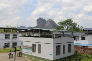 Bir Çin kırsal köyünün okulunda, tepesinde güneş panelleri ve yeşil çatı kiremitleri olan açık hava çatısı vardır. Önünde iki farklı yükseklikte beyaz bina, orta büyüklükte bir yapının yanında küçük bir bina bulunur..