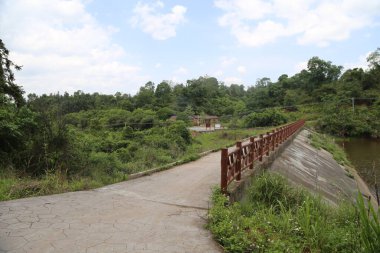 Yeşil bitki örtüsü ve her iki taraftaki uzak evler ile çevrili küçük bir köprü baraja gider. Kamera yer seviyesine yakın bir açıyla konumlandırılmış ve tüm manzarayı gösteriyor..