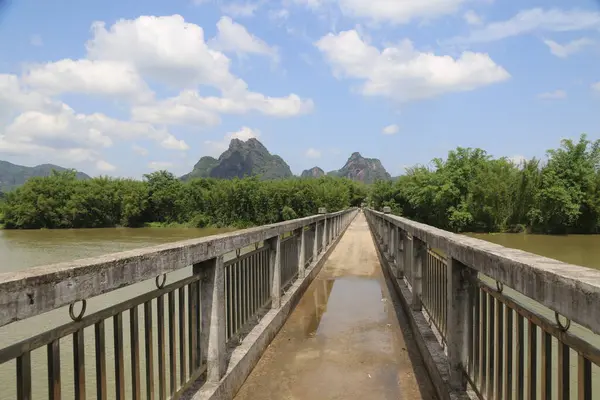 Nehrin üzerinde yeşil bitki örtüsü ve uzak arkaplanda dağlar olan bir köprü. Gökyüzü mavi ve üzerinde beyaz bulutlar yüzüyor..