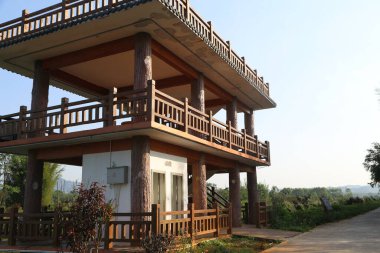 İkinci katında açık hava balkonu olan iki katlı ahşap bir bina. Tayland mimarisi tarzında dizayn edilmiş ve yemyeşil ile çevrili. Kırsal bir evin önünde manzarada geniş açılı mercek ve sıcak ışıklandırma yer alıyor.