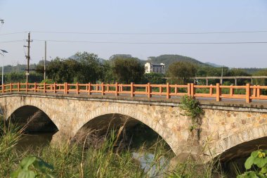 Çin kırsal bölgelerindeki nehir üzerindeki taş kemer köprüsü açık gökyüzü ve her iki taraftaki uzak dağlarla kırmızı ahşap korkuluklardan yapılmıştır. Kompozisyon ahşap korkuluklara odaklanıyor ve yanında yeşil otlar var..