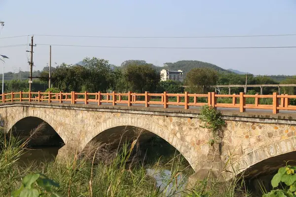 Çin kırsal bölgelerindeki nehir üzerindeki taş kemer köprüsü açık gökyüzü ve her iki taraftaki uzak dağlarla kırmızı ahşap korkuluklardan yapılmıştır. Kompozisyon ahşap korkuluklara odaklanıyor ve yanında yeşil otlar var..