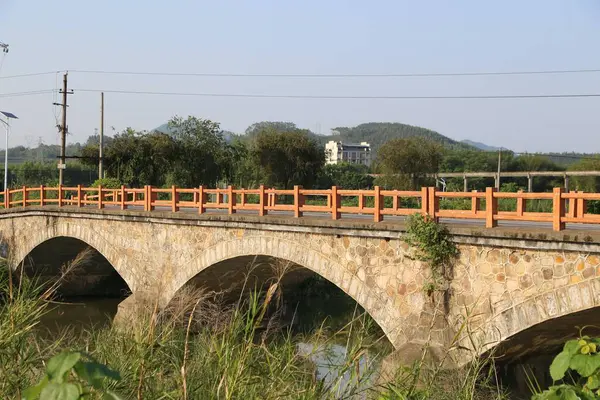 Çin kırsal bölgelerindeki nehir üzerindeki taş kemer köprüsü açık gökyüzü ve her iki taraftaki uzak dağlarla kırmızı ahşap korkuluklardan yapılmıştır. Kompozisyon ahşap korkuluklara odaklanıyor ve yanında yeşil otlar var..