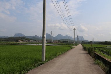 Uzakta, Çin 'in güneyindeki pirinç tarlalarına ve yeşil dağlara giden asfalt bir yol var. Uzak dağ zirveleri mavi gökyüzünün altında belli belirsiz görünür..