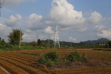 Sahada, önünde bir elektrik direği ve arkasında da iki beyaz metal elektrik hattı kulesi var. Çin 'in kırsal kesimlerinde bulutlu mavi bir gökyüzü görülebilir ve resmin bir tarafında küçük bir köy görülebilir..