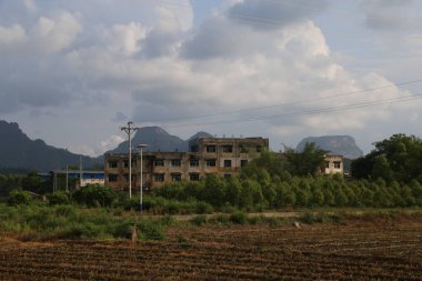 Tarlalarla çevrili harap bir bina, arka planda dağlar ve bir tarafında elektrik direği olan. Fotoğraf gündüz vakti pencerenin dışından çekildi..