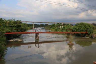 Nehir akıyor ve üzerinde fotoğrafın sol tarafında hiç insan olmayan eski bir kırmızı metal köprü var. Fotoğrafın bir tarafında yeşil bitkiler olmalı, elektrik hatları görünürde.