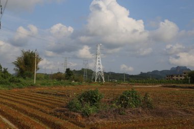 Sahada, önünde bir elektrik direği ve arkasında da iki beyaz metal elektrik hattı kulesi var. Çin 'in kırsal kesimlerinde bulutlu mavi bir gökyüzü görülebilir ve resmin bir tarafında küçük bir köy görülebilir..