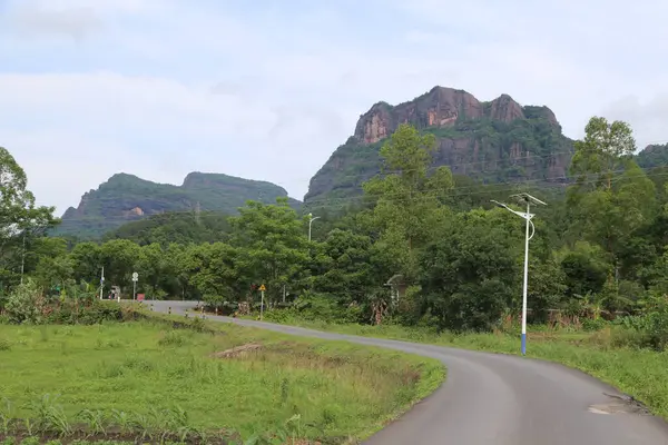 Yol dağ zirvesine çıkar, her iki tarafında yeşil ağaçlar ve çimenler vardır. Uzak manzara, yoğun ormanlarla kaplı iki yüksek dağın önünde eski kırmızı bir uçurum olduğunu gösteriyor..