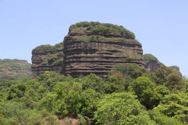 Tepesinde yeşil orman olan bir dağ. Dağ çok yüksektir ve kayalık bir yüzeyi vardır.