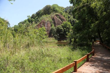 Ahşap bir geçit, yoğun bitki örtüsü ve çayırlarla çevrili kırmızı bir kaya dağının eteğine çıkar. Önünde altında su olan açık bir göl var..