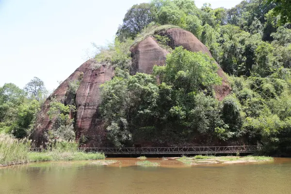 Ormanın ortasında, üzerinde demir bir köprü ve altında küçük bir göl olan kırmızı bir kaya dağı. Fotoğraf arkadan çekilmiş, kayaların tepesinde ağaçlar büyümüş..
