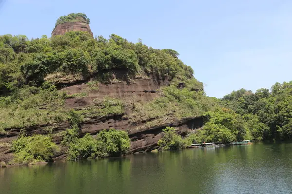 Bir dağın yüksek dağı, her iki tarafında yeşil bitki örtüsü ve altında berrak su ile. Ayağında çok uzaklardan görülebilen antik bir taş tekne var.