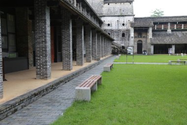 Eski Çin kalesinin avlusunda taş tuğlalar, gri tuğlalar ve her iki tarafta yeşil çimenler bulunur. Önünde açık bir alan var, yanında banklar var..